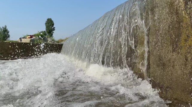 Artificial concrete check dam directs water into churning, frothy river stream lined with green trees