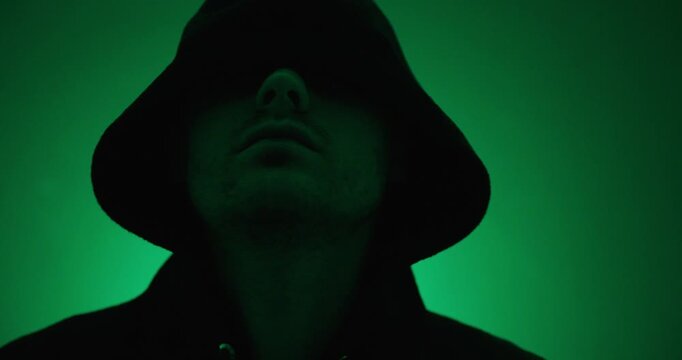 Hooded man walking slowly toward the camera under green lighting. Dramatic concept of anonymity, mystery, cybersecurity, suspense, and hidden identity.