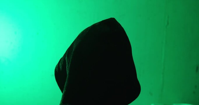 Rear view of a hooded person standing under green lighting. Dramatic concept of anonymity, secrecy, cybersecurity, mystery, and hidden identity.