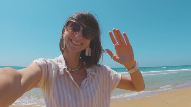A smiling woman wearing dark sunglasses and a striped blouse smiles and waves at the camera, enjoying the warm sun and the breeze on the beach.