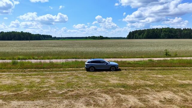 Blue SUV parked alongside a vast golden wheat field under bright blue skies, aerial view