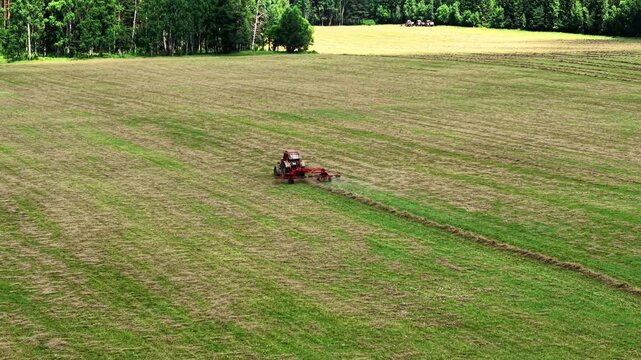 Red tractor mowing wide green field with freshly cut grass and trees in the background, aerial view
