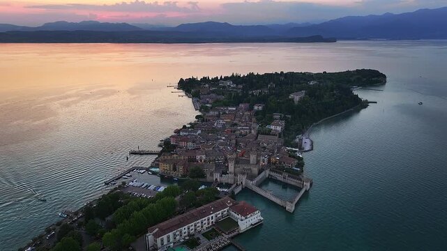 Sirmione, Italy - 4K Aerial view of the comune of Sirmione with Scaligero Castle (Castello Scaligero di Sirmione), located at the lower part of Lake Garda. Sunset, dramatic golden sky and clouds