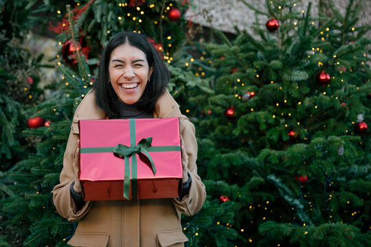 Smiling woman holding a red Christmas gift outdoors by a tree