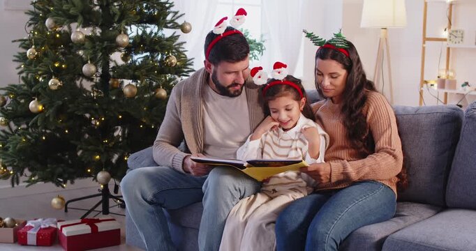 Parents and child girl reading a holiday book. At home by festive Christmas tree, mother and father guide daughter as family gifts and lights make warm atmosphere. Winter holidays celebration. 