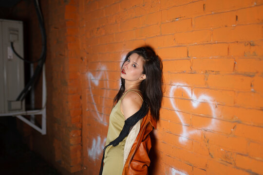 Model in raincoat leaning on orange brick wall with graffiti hearts