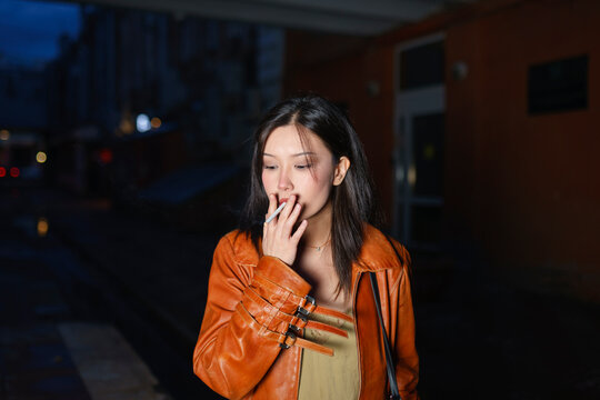 Smoking woman in a raincoat holds a cigarette in the city at evening