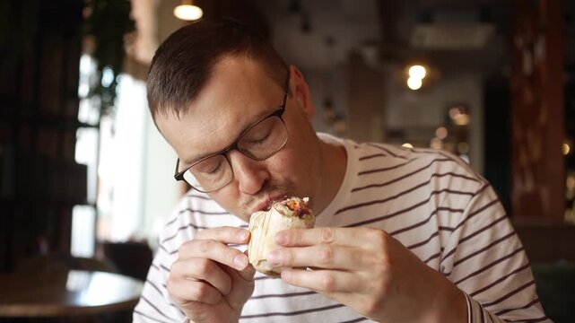 Young man in glasses enjoying a delicious vegan falafel roll at a cafe table, taking a large bite and chewing the healthy wrap with evident satisfaction during his lunch break