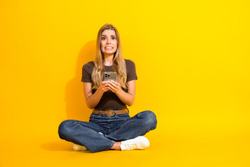 Young woman sitting cross-legged on a bright yellow background nervously holding a smartphone with...
