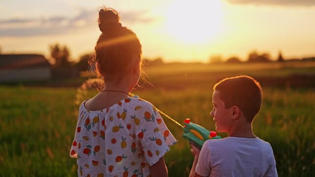Two children are standing in a field, spraying water from colorful guns as the sun sets. They enjoy the warm evening and the fun of water play