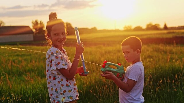 Two children are in a green field playing with water toys as the sun sets. They are spraying each other while laughing on a warm summer evening
