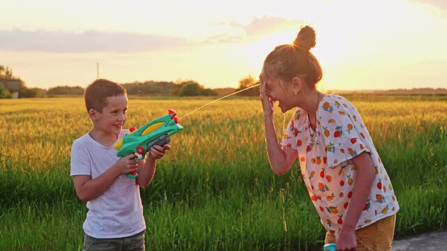 Two children enjoy playing with a water gun in a field. The sun sets in the background, creating a warm and lively atmosphere