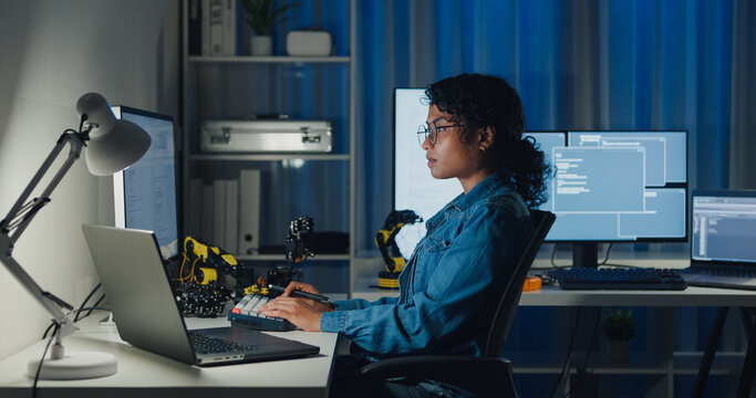 Young African American female programmer coding AI software on multiple screens in night office. Network security, cybersecurity, machine learning, working late, overtime, robotics automation concept.