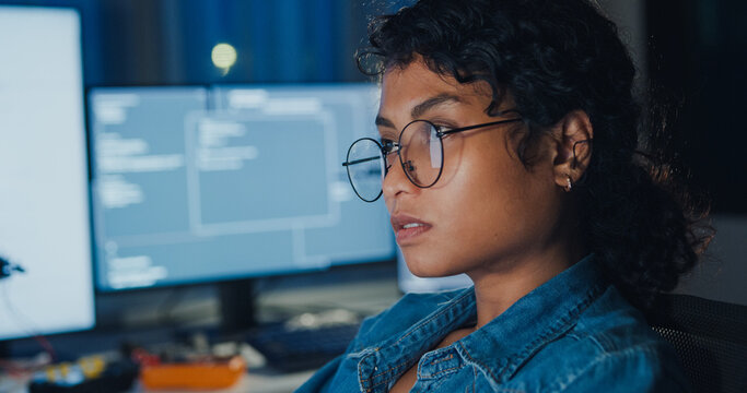 Close-up young Black female programmer coding AI software on multiple screens in night office. Network security, cybersecurity, machine learning, working late, overtime, robotics automation concept.