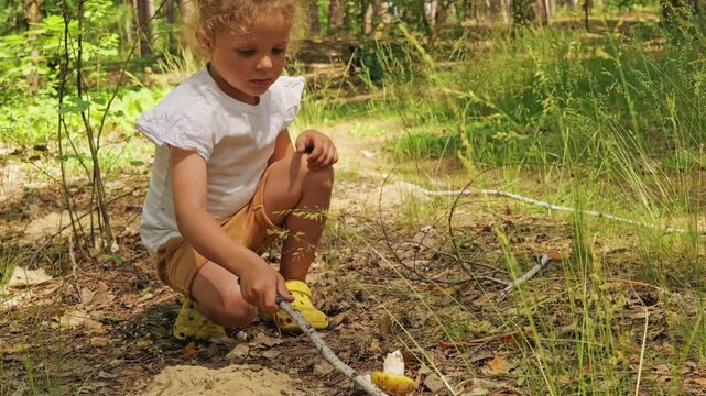 A child is digging in the forest floor using a stick. The child looks focused on finding something in the dirt. The sun shines through the trees