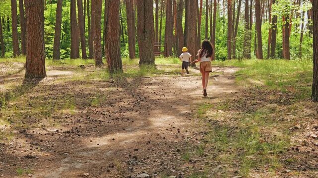 Two children enjoy a day in the forest. The girl runs with a stick in hand while the boy approaches her. They seem happy and engaged in play