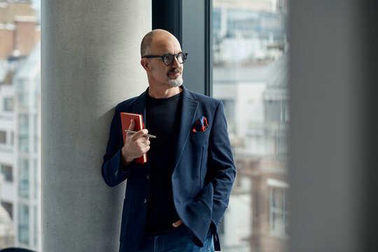 A businessman stands by an office window holding a notebook in London