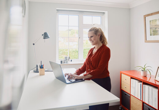 A woman types on a laptop at a standing desk in a home office