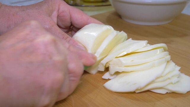Several even slices of scamorza cheese are cut in succession on a wooden board, methodically preparing enough cheese to generously layer a traditional southern Italian baked pasta dish.