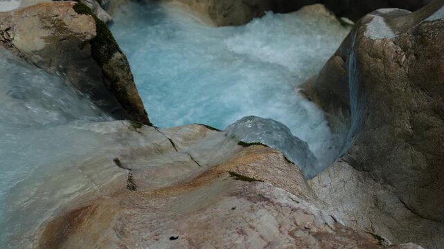 Close-up view of pale blue water rushing through smooth rock formations in a narrow mountain stream. Small patches of moss grow along the wet stone edges.