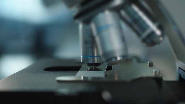 Gloved lab worker moves the microscope objective over a prepared slide during pharmaceutical quality control testing.