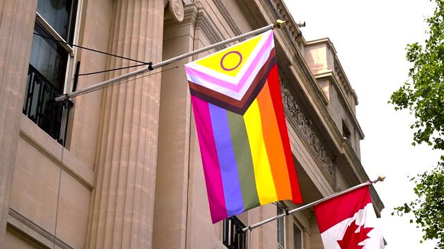 Progress Pride flag and Canadian flag move in the breeze outside Oceanic House against a stone fa&ccedil;ade and summer sky.