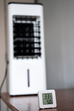 A digital room thermometer displaying a high temperature of 33&deg;C, set against the blurred background of a portable air cooler. Summer heat and the concept of climate control