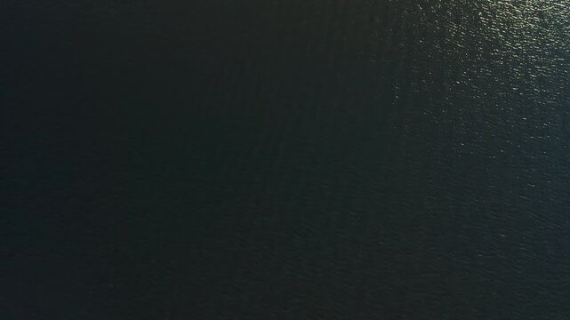 Subtle ripples move under a patch of bright sun glint on an otherwise dark, calm water surface seen from high altitude.