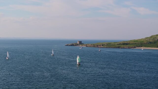 Small sailboats with colourful spinnakers move across open sea toward a Martello tower on a green Irish headland.