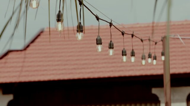 Description: String of hanging bulb lights strung against a terracotta tiled roof and pale sky at a garden venue