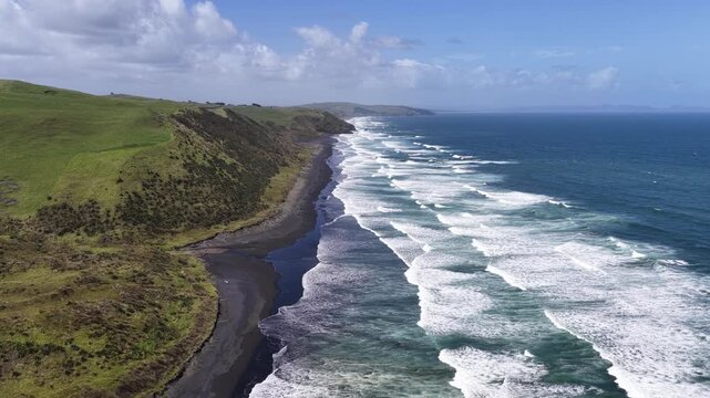 Wide aerial view following the dramatic black sand beach of New Zealand's North Island, where rolling ocean waves meet steep coastal hills in a spectacular natural seascape.