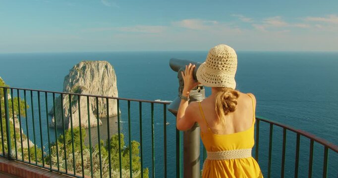 Traveler using public telescope to view Faraglioni sea stacks on coast in Capri Italy