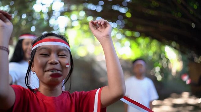 Smiling child in red shirt lifts both arms in playful celebratory pose, facing camera inside shaded tropical gathering space with blurred children behind, ribbon barrier, bright foliage, and warm