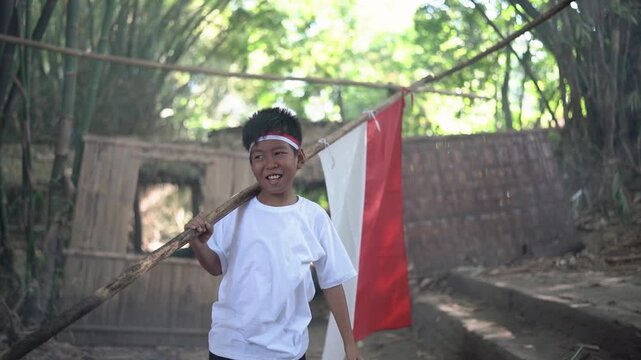 Young boy in rustic village setting leans playfully with flagpole across shoulder while extending one arm outward, smiling with joyful expression, red and white cloth hanging nearby, bamboo fencing