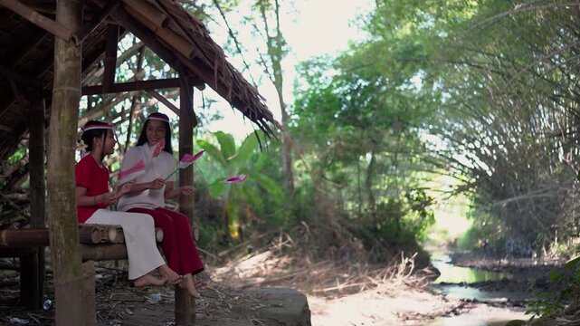 Two young girls sit beneath wooden shelter beside narrow creek in lush tropical forest, smiling and holding small pink and white flags while natural light filters through dense greenery