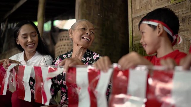 Elderly woman smiles while seated with young woman and child inside rustic bamboo home, with child turned toward her behind red and white ribbon garland, creating warm candid family scene