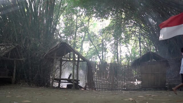 People walk past bamboo huts and rustic fence at rural village entrance beneath dense trees, with red white flag, bright backlit haze, soft shadows, weathered wood textures, and open dirt ground