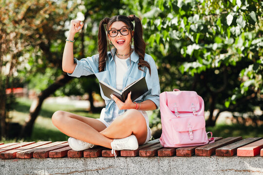 Young woman student outdoors smiles while reading a notebook beside a pink backpack in a sunny park enjoying study break and casual fashion