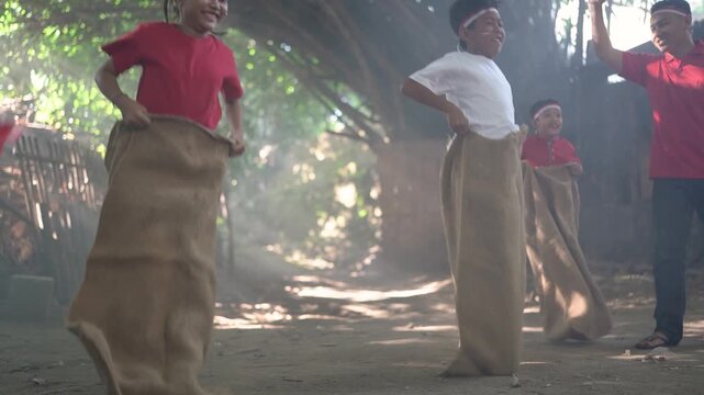 Children hop inside burlap sacks during playful rural race while adult signals beside dusty path, with bright red shirts, white headbands, rustic fence, warm haze, and trees, with children, hopping