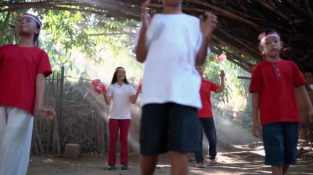 Child moves across foreground while group stays behind in rustic village yard, wearing red, white, and black clothes with matching headbands, beside bamboo fence, thatched roof, dusty ground, and