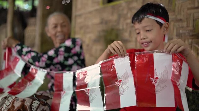 Cheerful boy in red shirt holds glossy red white bunting across foreground inside rustic woven wooden interior, while smiling older woman sits behind in soft focus under warm light, creating festive