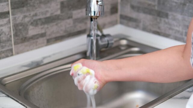 Hand rinsing a kitchen sponge under running water in a stainless steel sink