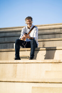 Portrait of smiling mature man sitting on steps with headphones and cell phone