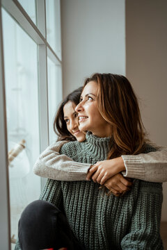 Happy mother and daughter looking through window while day dreaming together at home