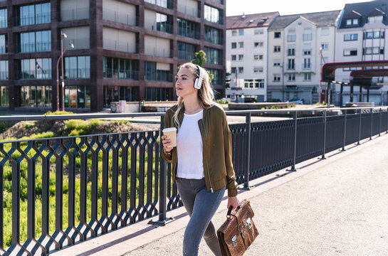 Young woman walking on bridge, drinking coffee, listening music with headphones