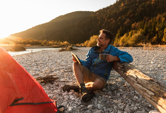 Mature man camping at riverside, holding tablet and cup