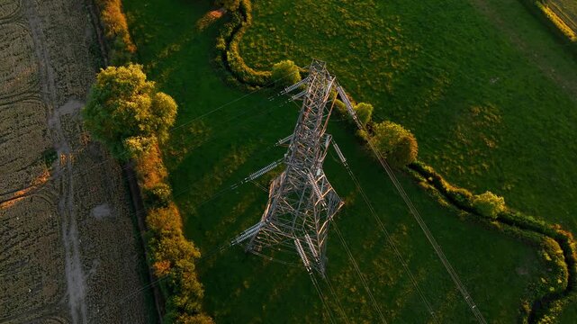 High voltage transmission tower above agricultural landscape at golden hour. Aerial view with power lines crop fields green meadows and rural infrastructure. Electricity distribution network energy