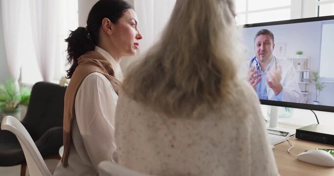 Senior woman doctor telemedicine video call online. At home a caregiver sits with patient as a doctor on the computer gives an online consultation. Trusted digital healthcare for aging families.