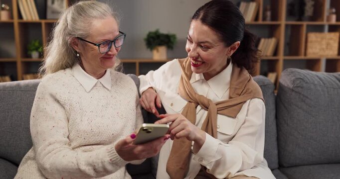 Senior caregiver helps woman with smartphone learning assistance. They sit on a sofa at home as the caregiver guides setup and apps. Clear concept of family technology support and learning.
