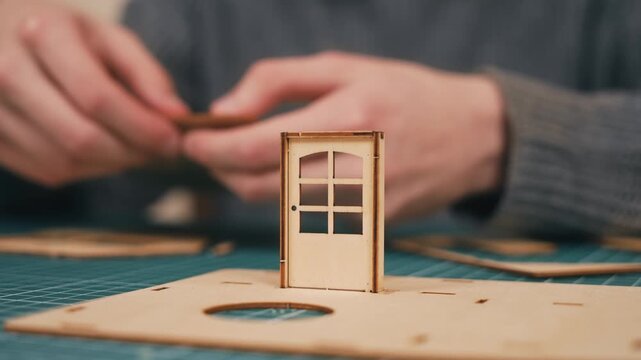 Assembling small wooden model house components behind completed front door. DIY miniature architecture crafting featuring detailed plywood construction and creative hobby process. Residential concept